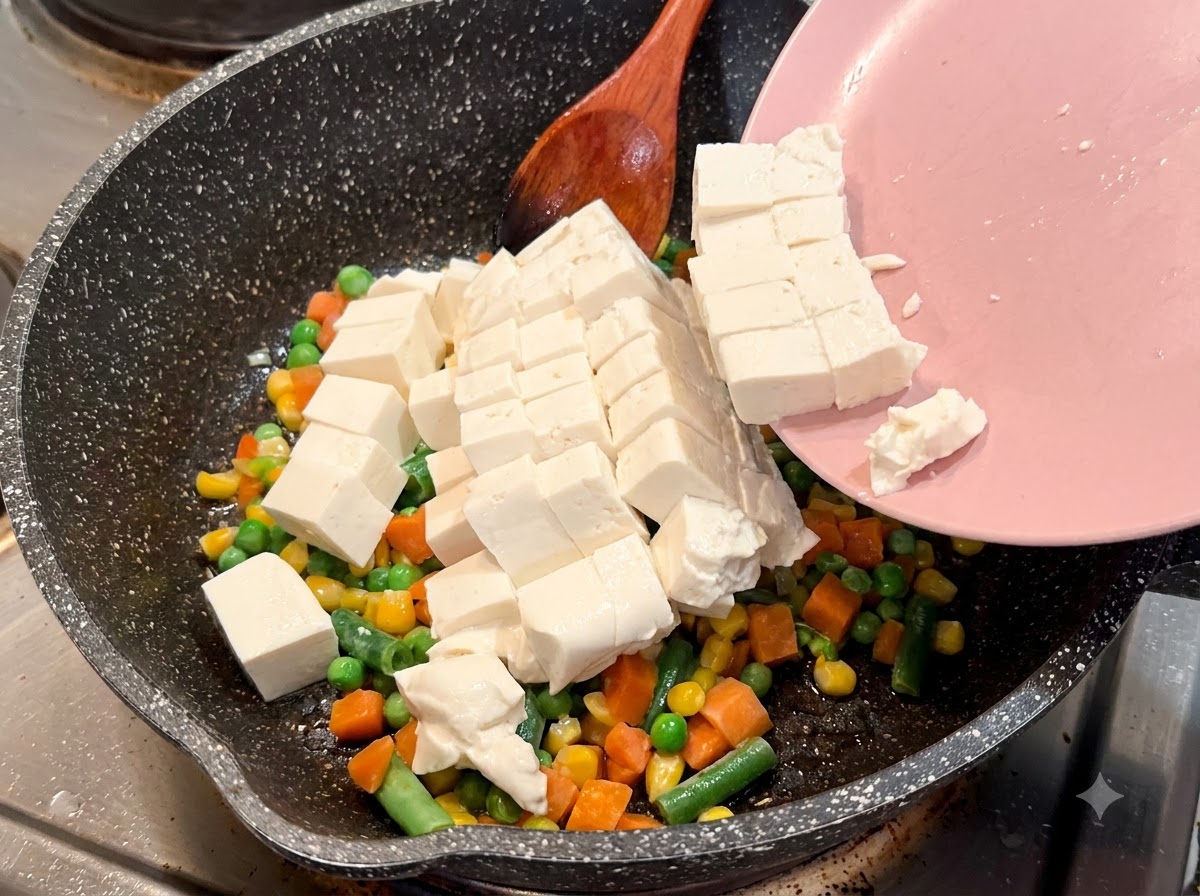 Tofu soyeux coupé en cubes glissant dune assiette rose vers une poêle mouchetée avec des légumes mélangés.