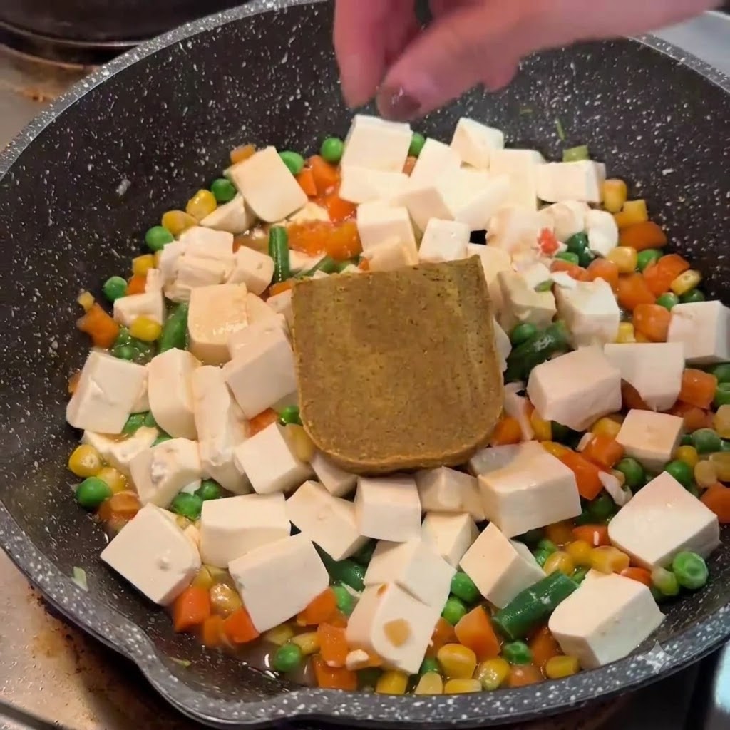 Une main plaçant un bloc de roux de curry brun solide sur le tofu et les légumes dans une poêle.