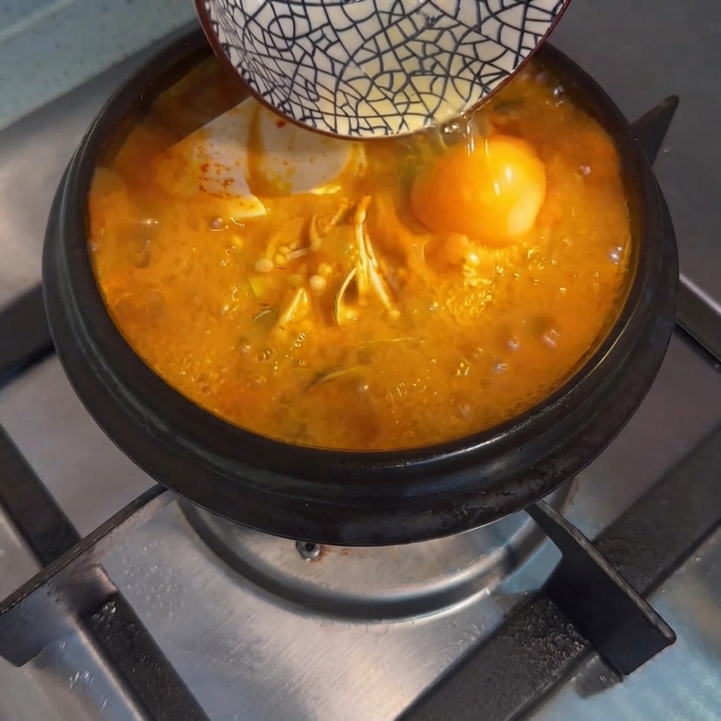 Un œuf cru versé dun petit bol dans le ragoût de tofu coréen bouillant.