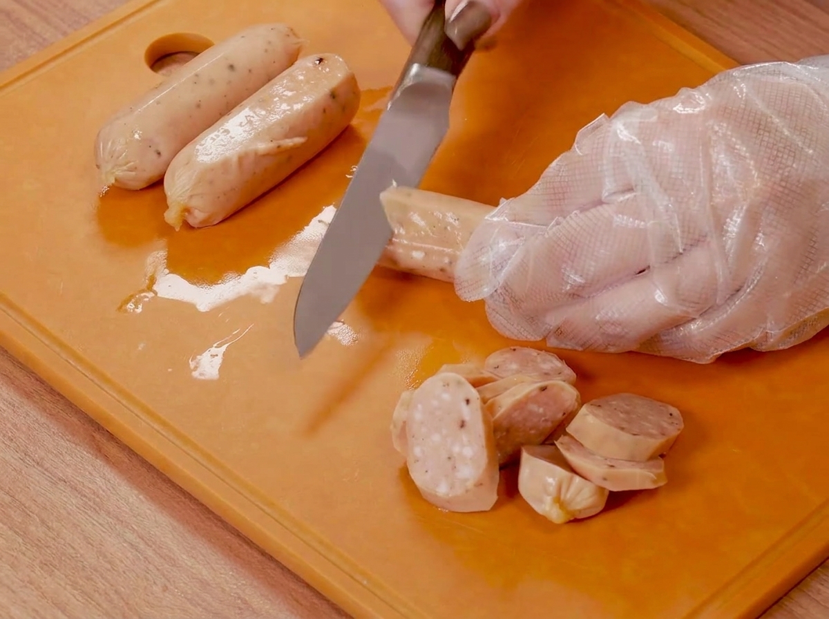 Mains gantées de plastique utilisant un couteau pour entailler de petites saucisses sur une planche à découper orange.