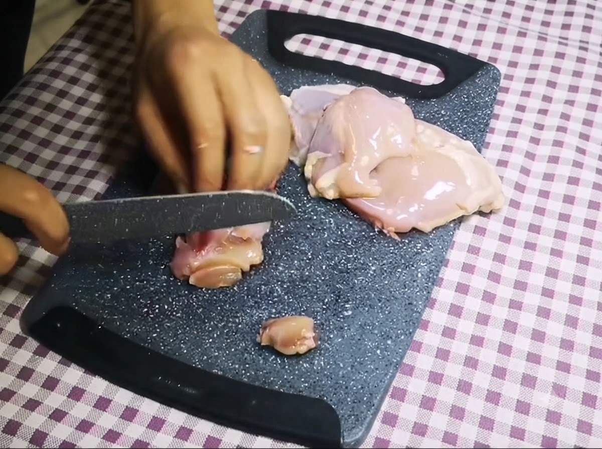 Un couteau tranchant du poulet cru désossé en petits morceaux sur une planche à découper grise.