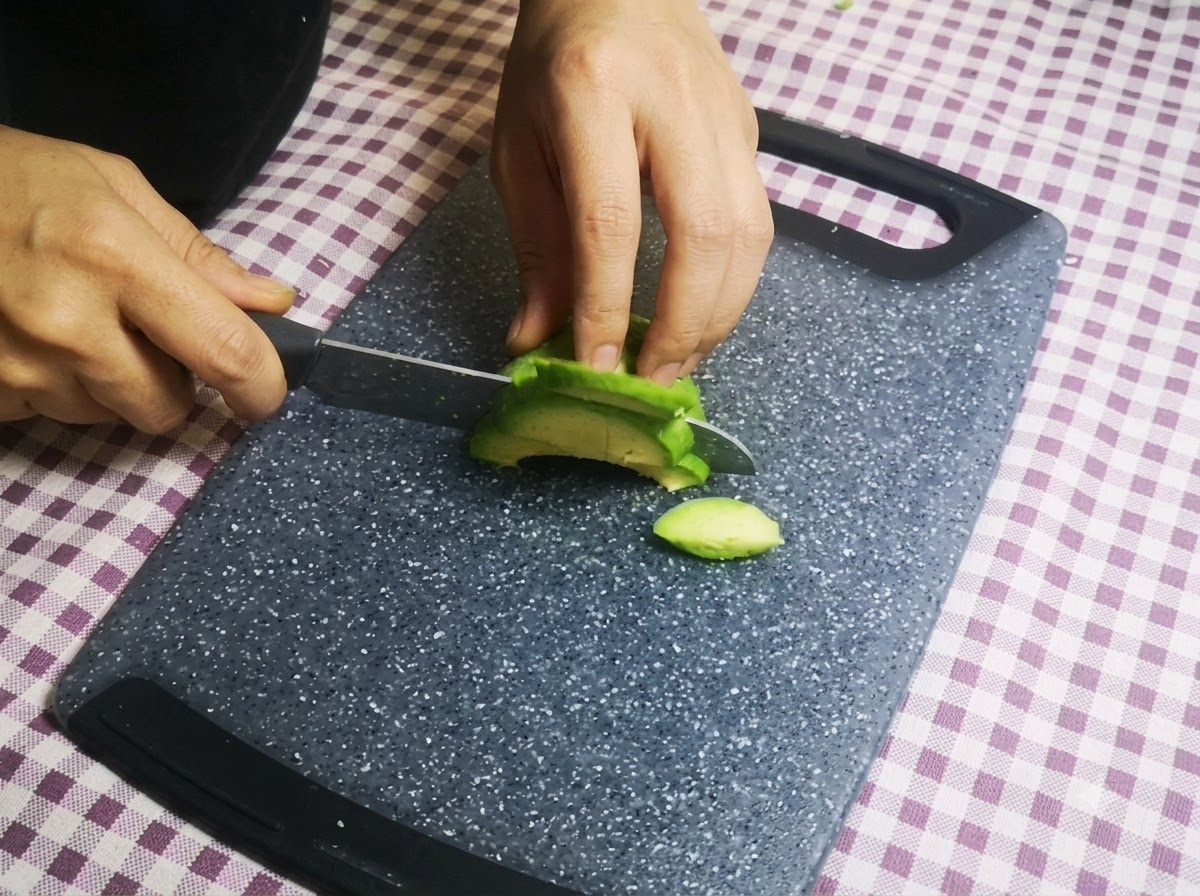 Des mains coupant un demi-avocat vert en fines tranches avec un couteau sur une planche à découper.
