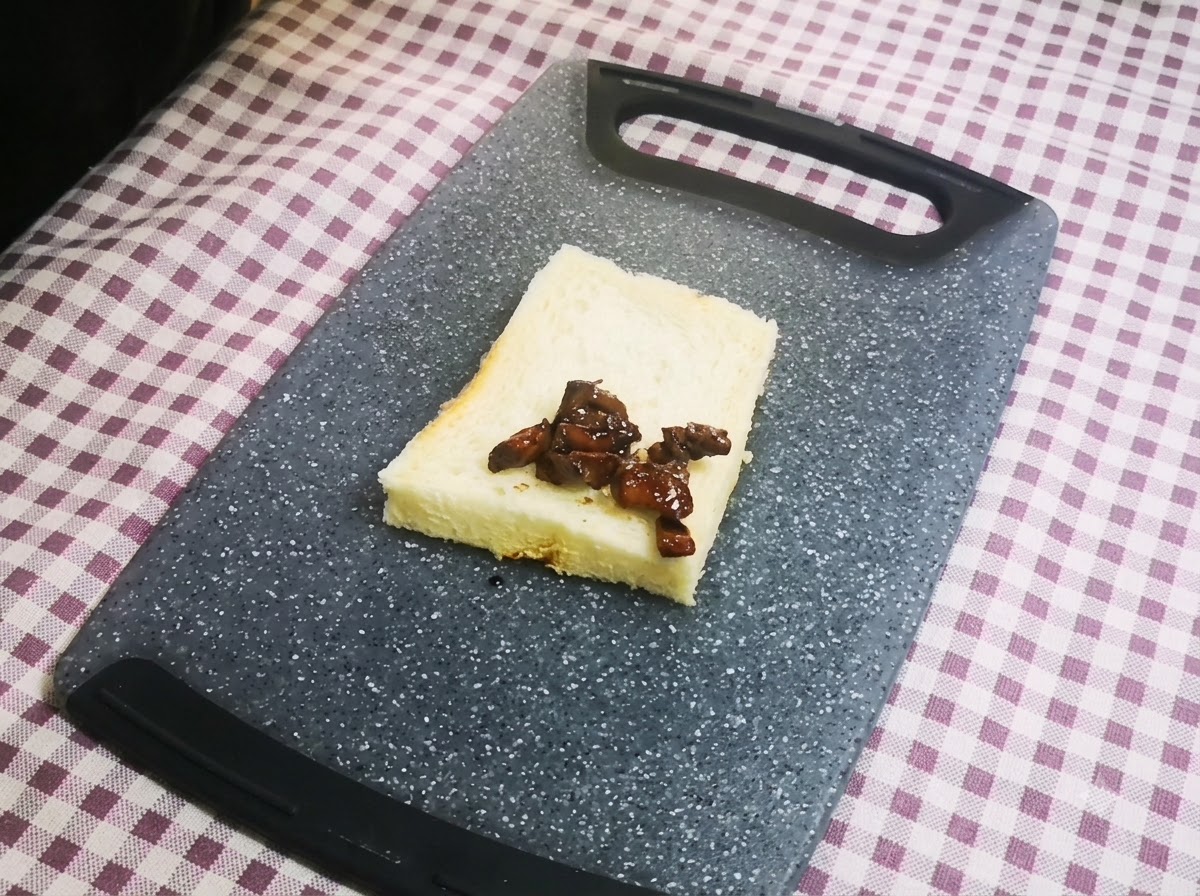 Un gros plan de morceaux de poulet nappés de sauce foncée disposés soigneusement sur une seule tranche de pain de mie posée sur une planche à découper grise.