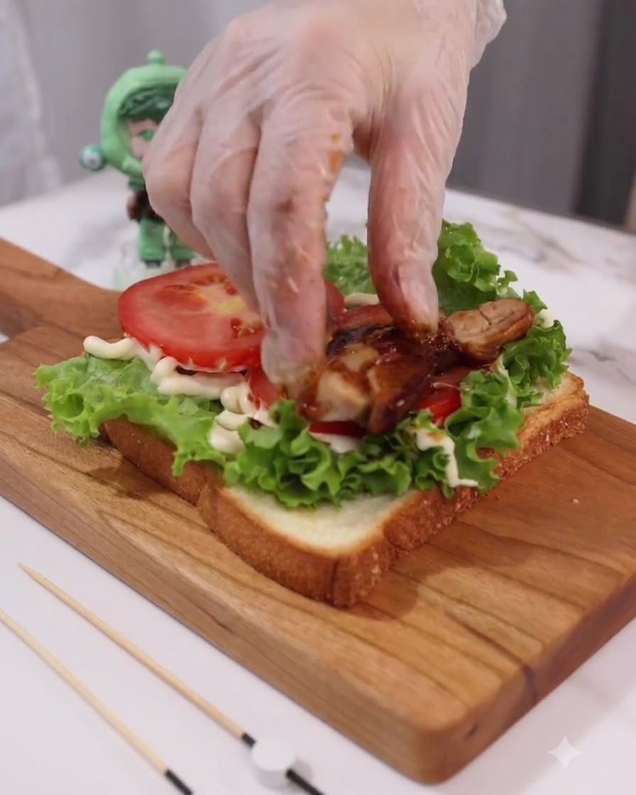 Main gantée plaçant des morceaux de poulet teriyaki cuits sur les couches de tomates et de laitue.