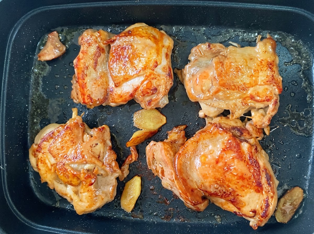 Quatre cuisses de poulet en train de frire dans une poêle avec une peau croustillante dorée et des tranches de gingembre.