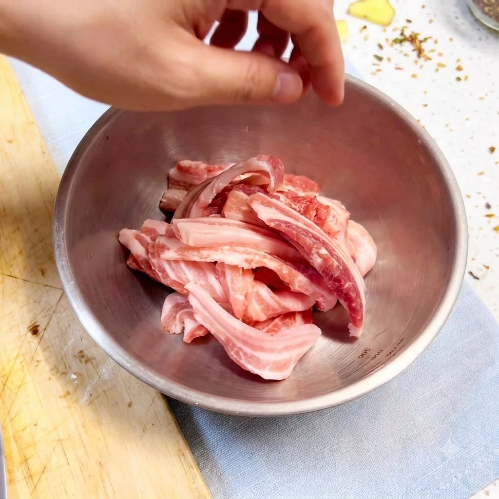Main gantée mélangeant des tranches de poitrine de porc avec des assaisonnements et des aromates dans un bol en métal.
