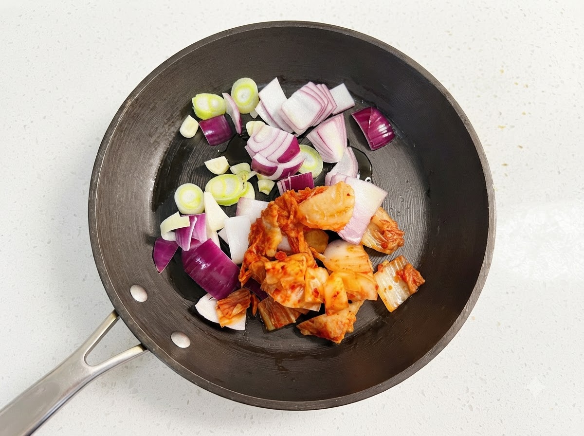 Tranches doignon violet, rondelles doignon vert et kimchi rouge haché grésillant dans une poêle noire avec de lhuile.