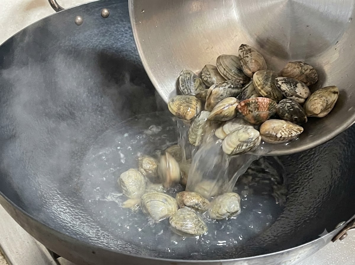 Des palourdes fraîches versées depuis un bol en métal dans un wok fumant.