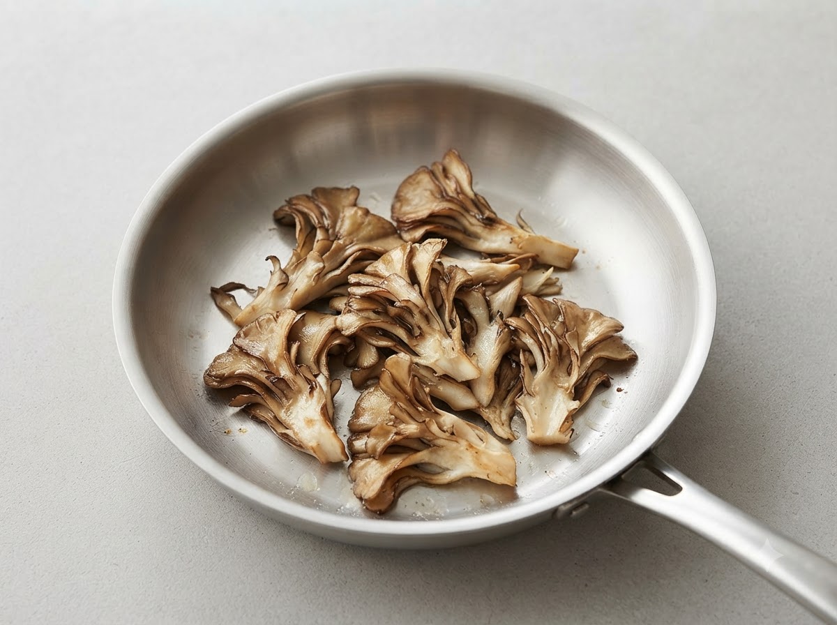 Des morceaux déchirés de champignons maitake en train de dorer dans une poêle en acier inoxydable propre.