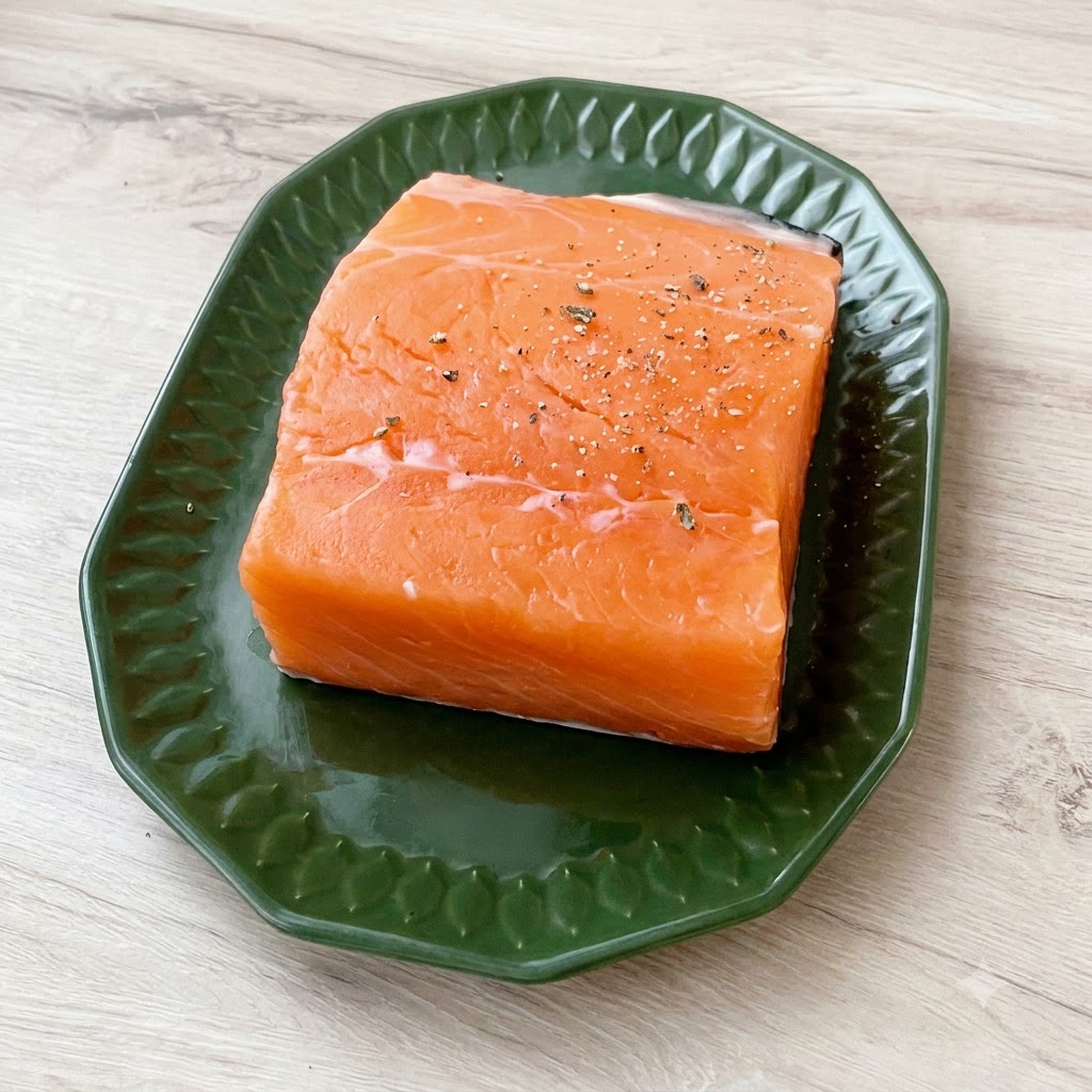 Un morceau épais de saumon frais posé sur une assiette verte, généreusement saupoudré de gros sel de mer et de poivre noir concassé.