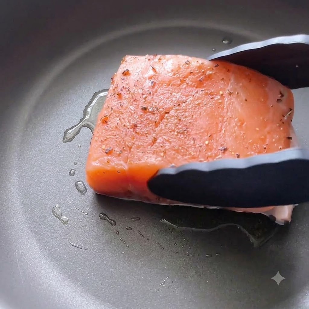 Un filet de saumon assaisonné déposé côté peau dans une poêle huilée à laide de pinces de cuisine noires.