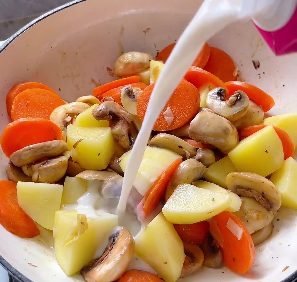 Du liquide versé depuis une brique rose dans une cocotte de cuisson remplie de cubes de pommes de terre, de rondelles de carotte et de champignons.