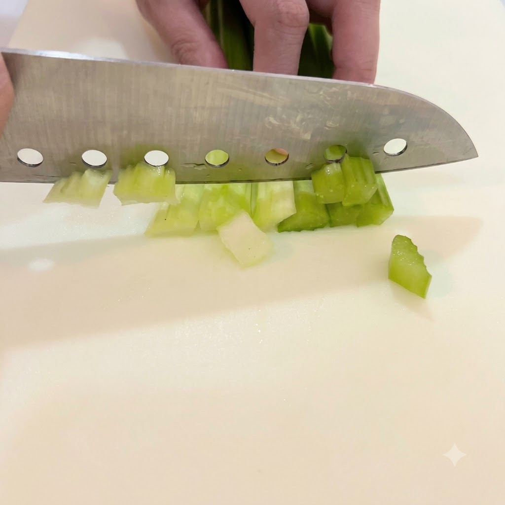 Une main guidant un grand couteau pour hacher des tiges de céleri vert vif en petits morceaux réguliers sur une planche à découper blanche.