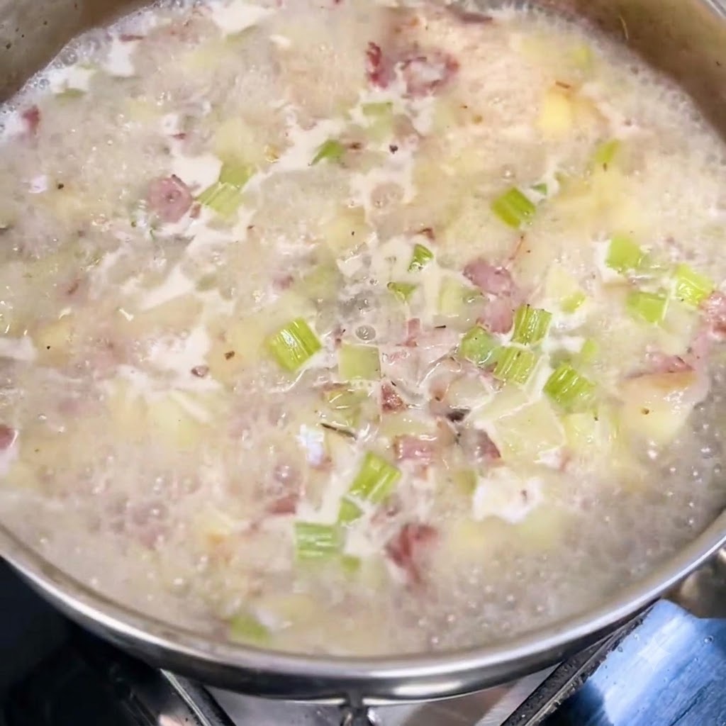 Lait et crème entière versés dans une casserole de soupe frémissante avec des morceaux visibles de pommes de terre, de céleri et de bacon.