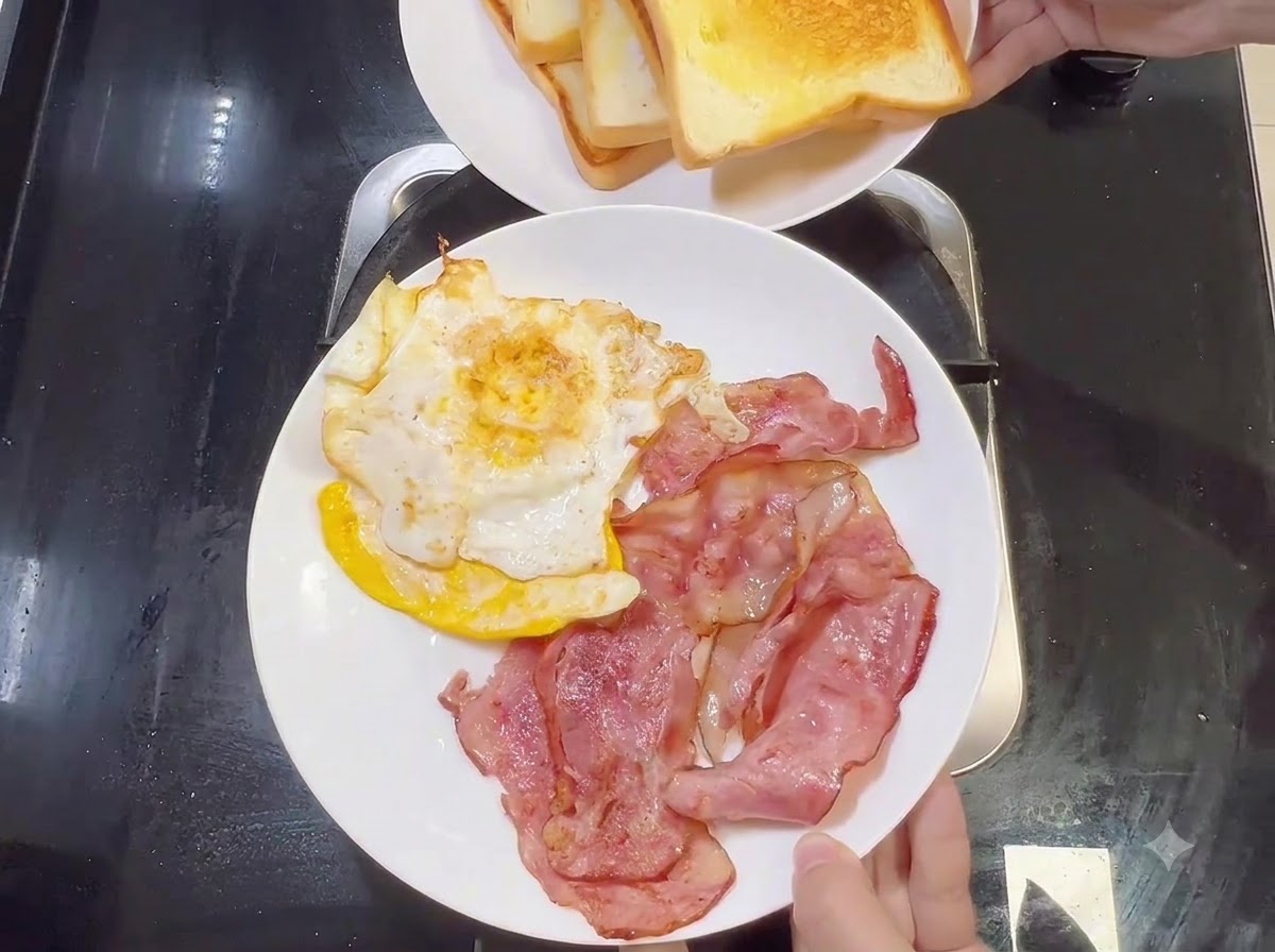 Une assiette blanche contenant des lanières de bacon cuites et des œufs frits, avec une autre assiette de pain toasté tenue au-dessus.