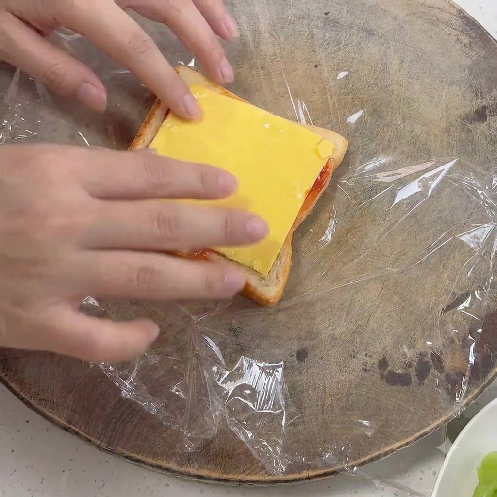 Des mains plaçant une tranche de fromage jaune carré sur un morceau de pain garni de sauce.
