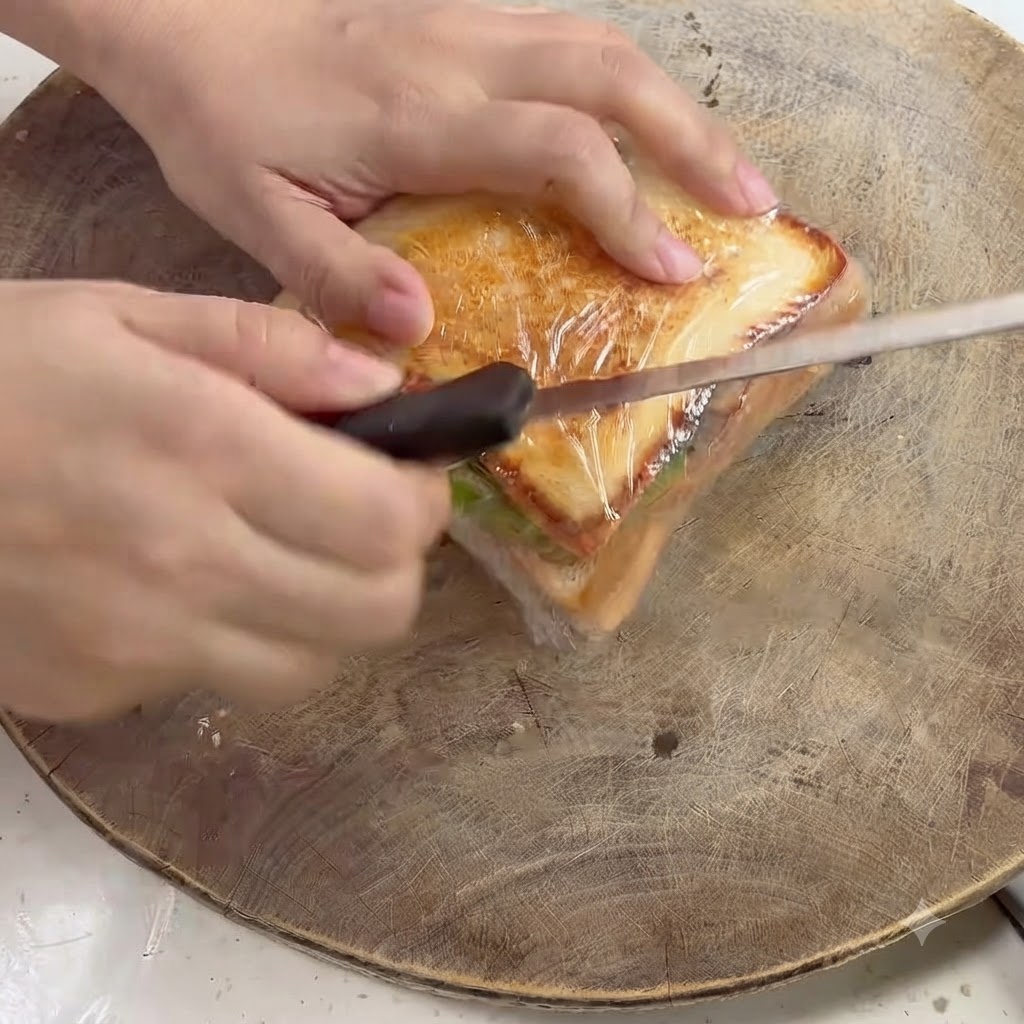 Un couteau de cuisine coupant en diagonale à travers un sandwich emballé dans du plastique sur une planche en bois.