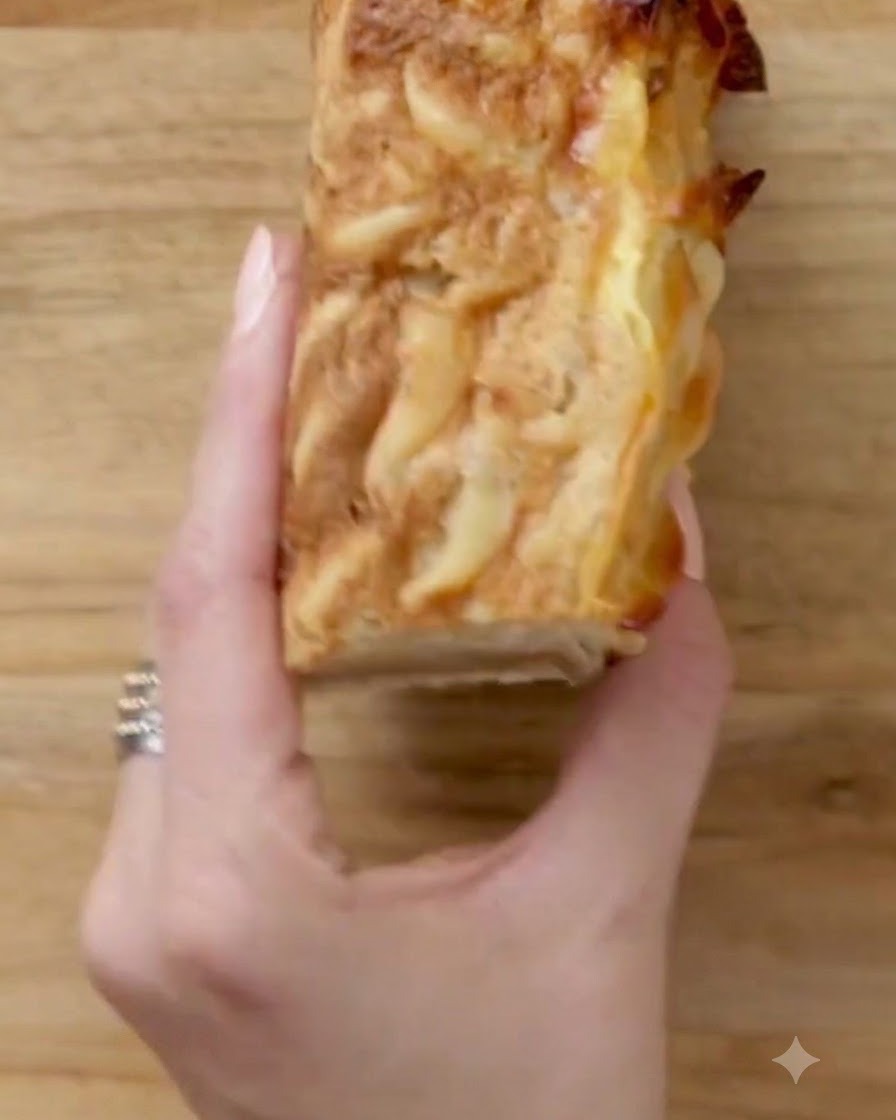 Une main tenant une portion carrée de kugel soigneusement coupée sur une planche en bois.