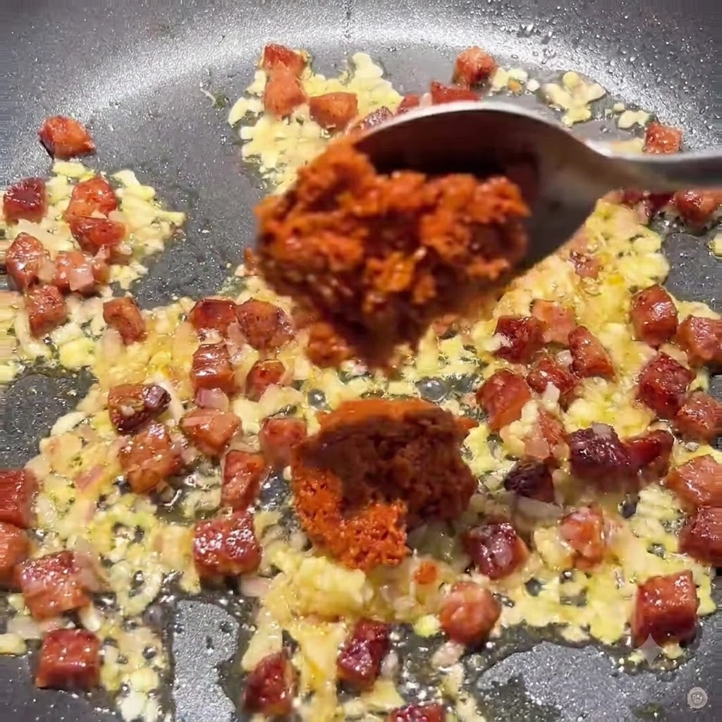 Une cuillère ajoutant de la pâte de tomates séchées épaisse et du concentré de tomate dans la poêle avec le mélange de chorizo et doignon.