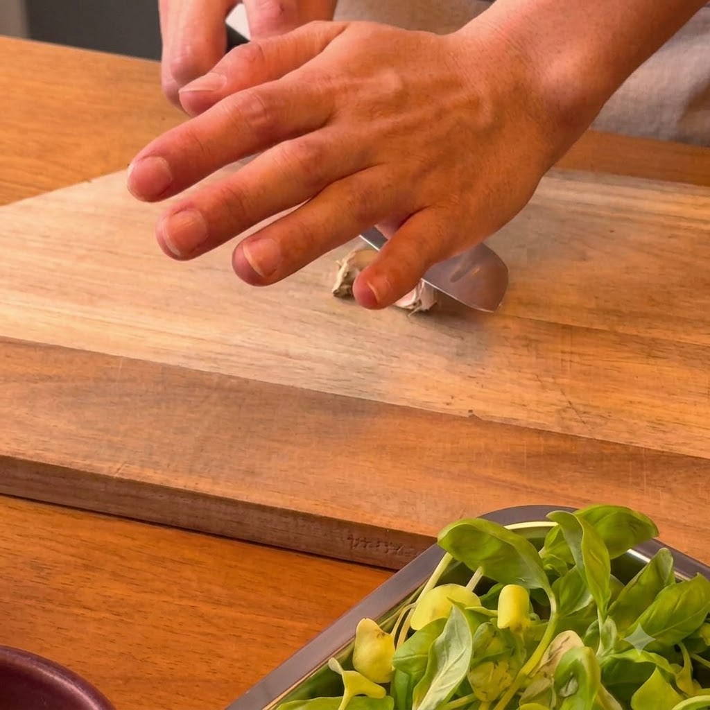 Une main pressant le côté plat dun couteau de chef sur une gousse dail posée sur une plancha en bois.