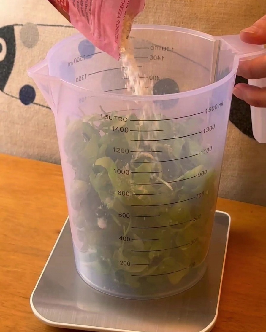 Du fromage râpé versé depuis un sachet rose dans un grand verre mesureur rempli de feuilles de basilic vert frais.