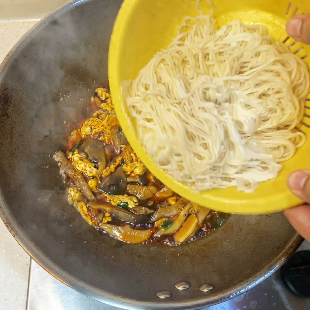 Des nouilles fines cuites versées dune passoire jaune dans le wok contenant la sauce et les ingrédients.