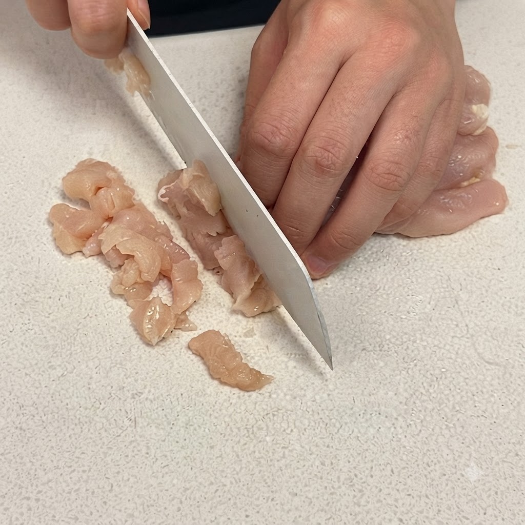 Gros plan sur un blanc de poulet découpé en petits cubes sur une planche à découper blanche avec un couteau de chef aiguisé.
