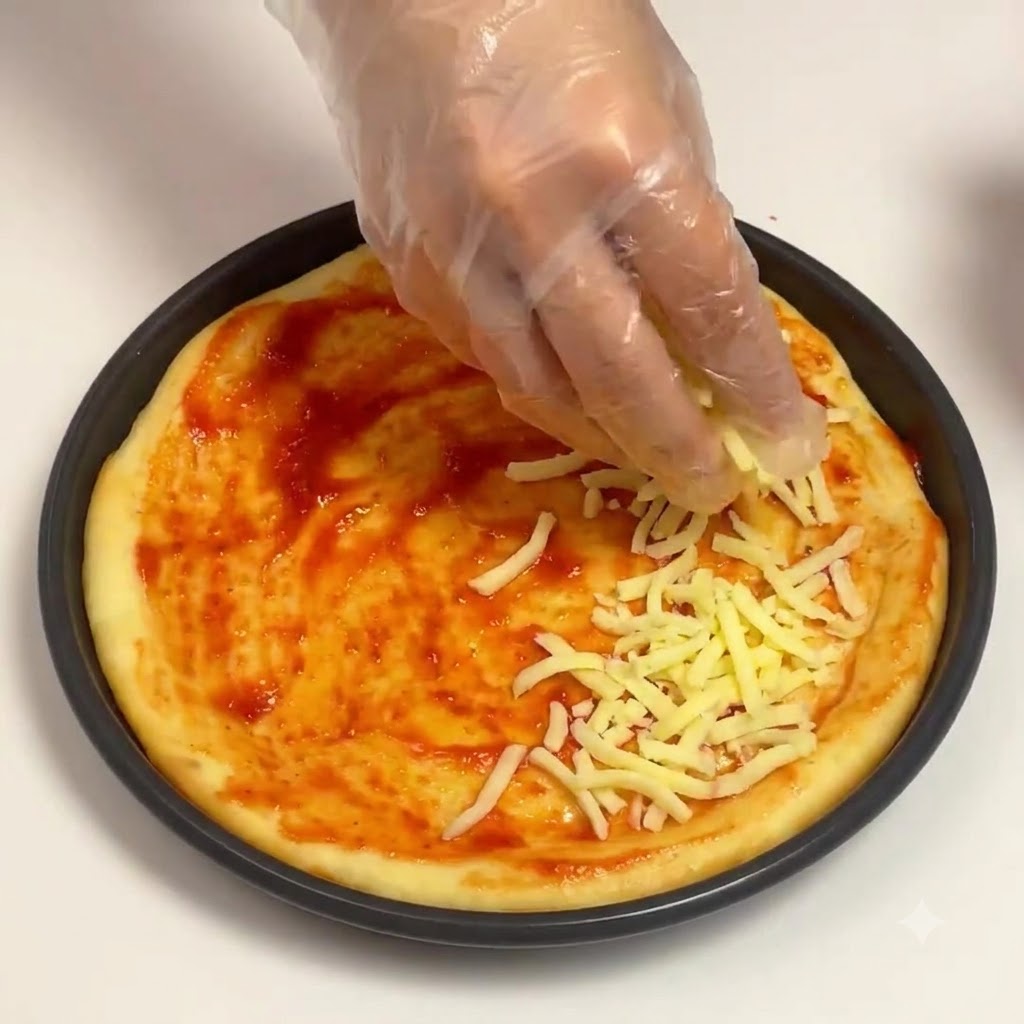 Une main gantée saupoudrant du fromage mozzarella râpé sur la base de sauce tomate dans une poêle à pizza ronde.