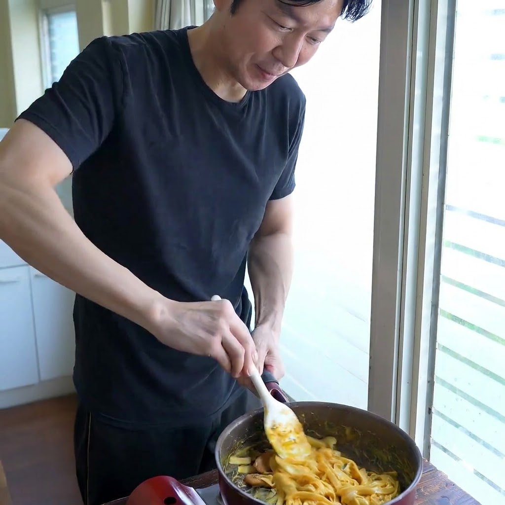 Une personne remuant des nouilles larges dans une poêle avec une spatule blanche tout en ajustant la consistance de la sauce.