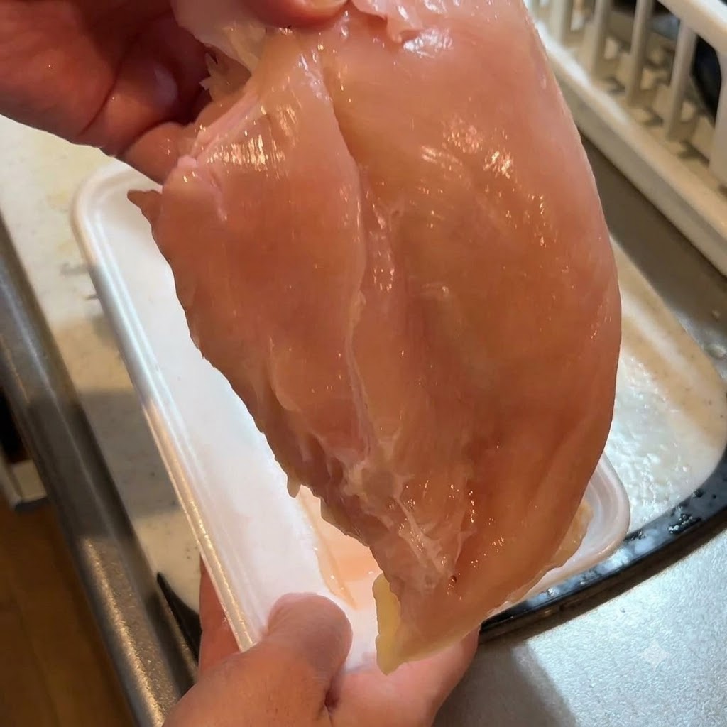 Un blanc de poulet cru tenu en main pendant que la peau est retirée avec un couteau.