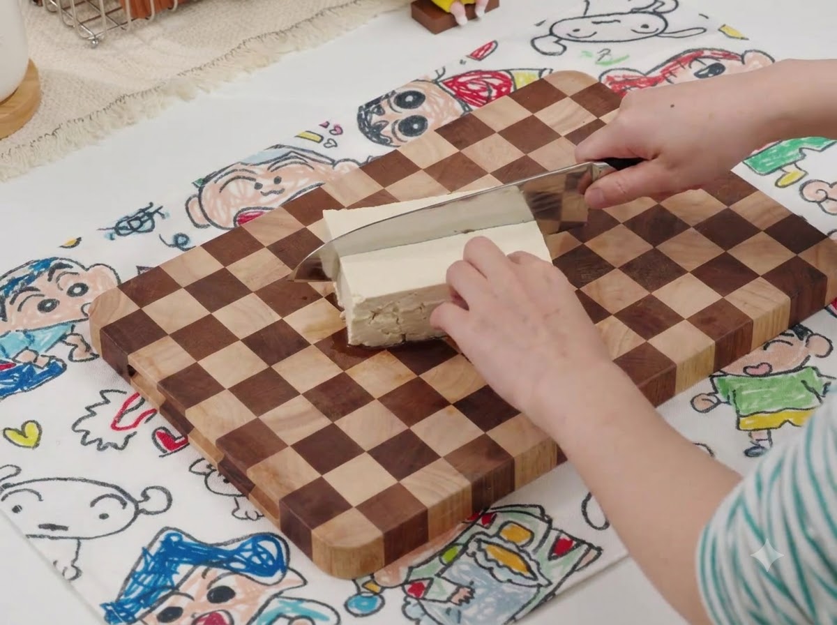 Un couteau coupant un bloc de tofu blanc sur une planche à découper en bois avec un tapis à motifs en dessous.