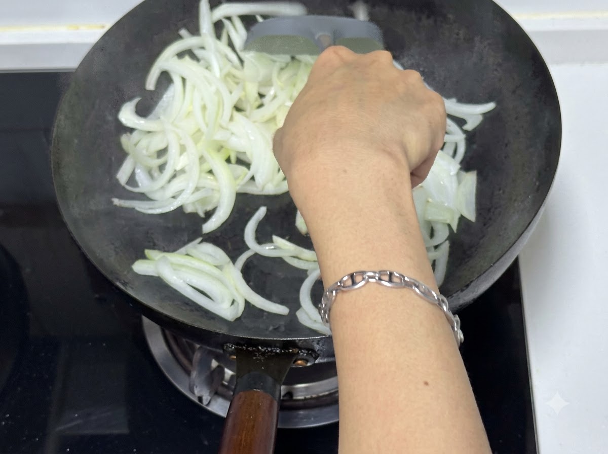 Une personne utilisant une spatule pour faire sauter des oignons émincés dans un wok, montrant des morceaux translucides et légèrement dorés.