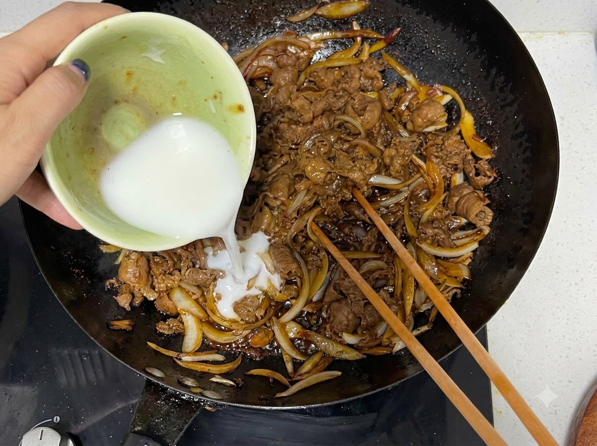 Un mélange de fécule de maïs versé dun bol dans un wok avec du bœuf et des oignons cuits, à côté de baguettes en bois.
