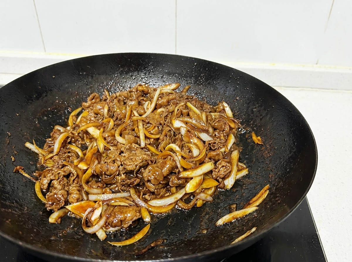 Gros plan sur des tranches de bœuf et des lamelles doignon sautées dans un wok noir avec des ustensiles de cuisine.