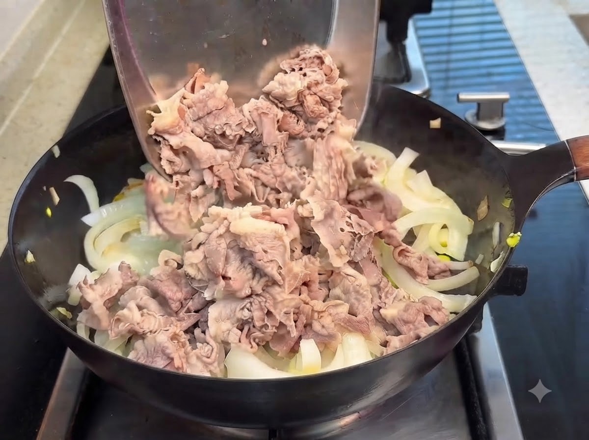 Des tranches de bœuf blanchies versées dune spatule en métal dans une poêle doignons translucides, avec de la sauce teriyaki versée par-dessus.