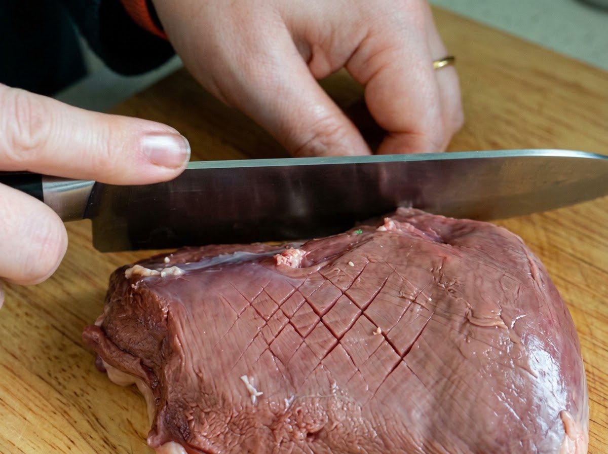 Un couteau de chef quadrillant la peau dun magret de canard cru en croisillons sur une planche en bois.