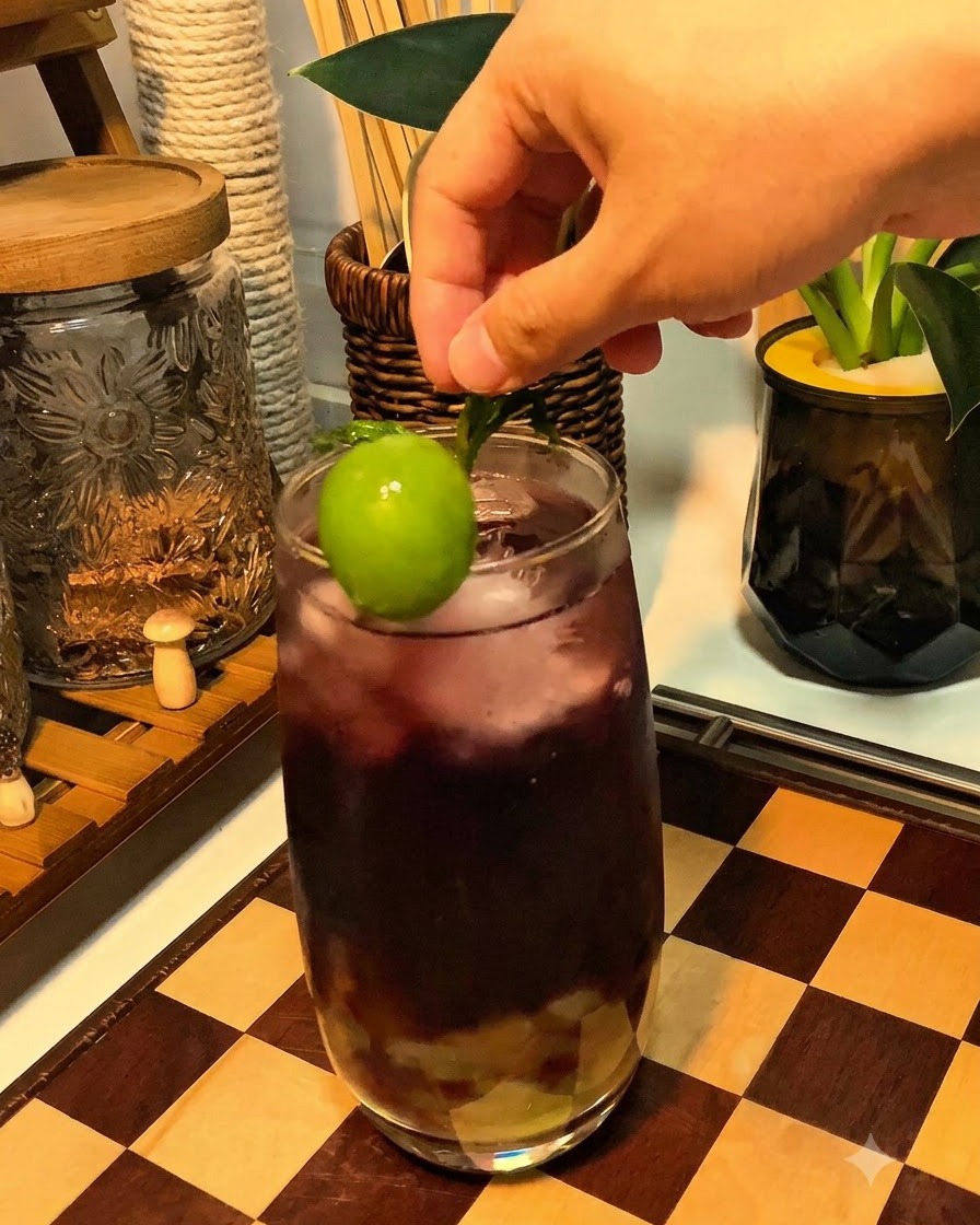 Une main plaçant un raisin vert frais et une garniture de menthe sur le bord dun verre de boisson pétillante pourpre.