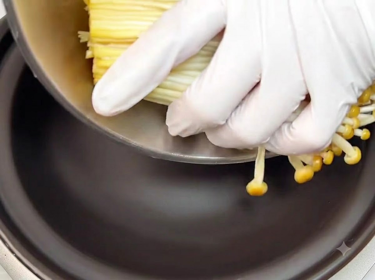 Mains gantées plaçant un bouquet de champignons enoki crus parés au fond dun plat en terre cuite noir.