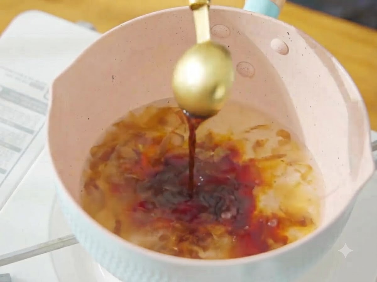 Verser de la sauce soja foncée dune cuillère dorée dans le bouillon de bonito frémissant de la casserole rose.