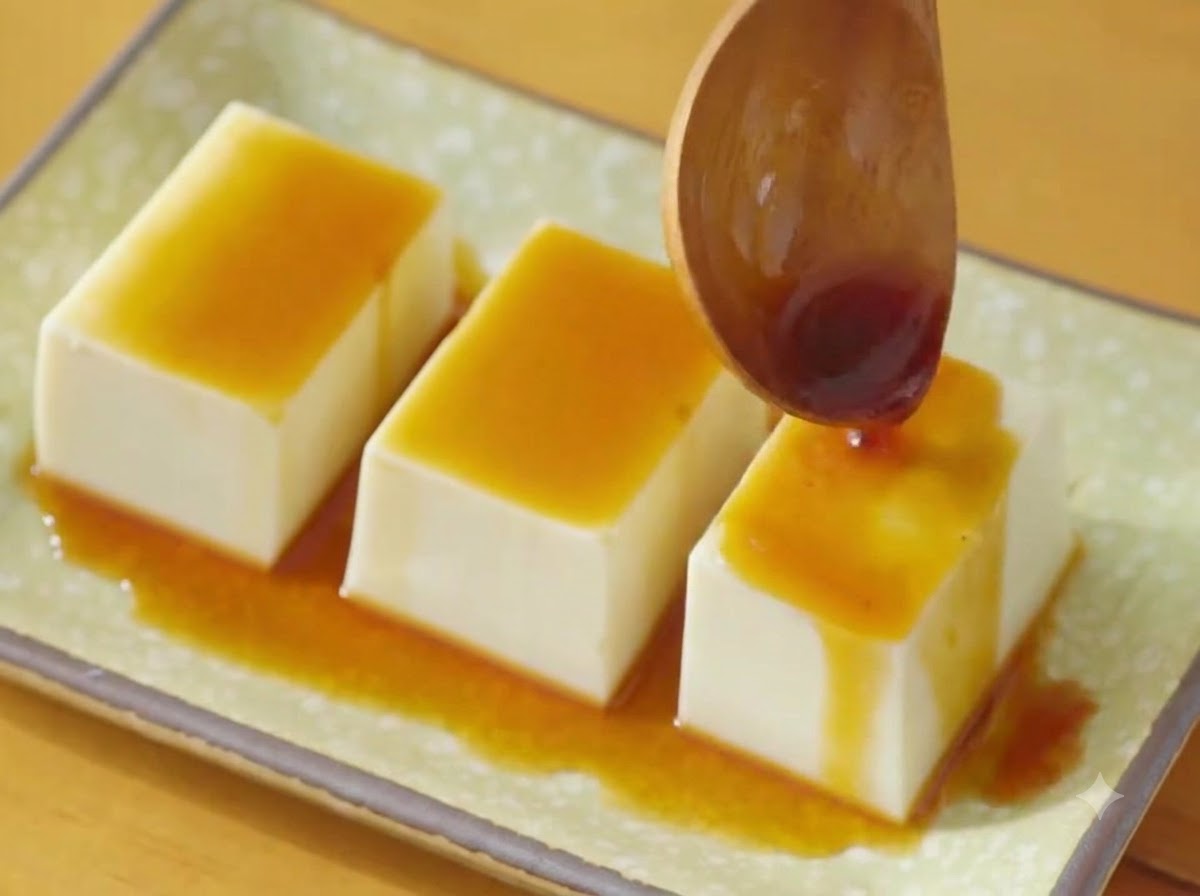 Une cuillère en bois versant de la sauce soja foncée sur trois rangées de tofu soyeux sur une assiette verte.