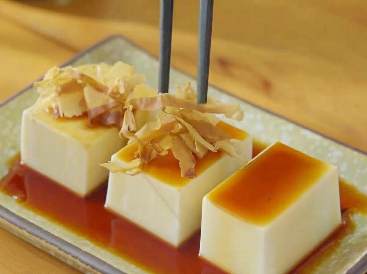 Des baguettes plaçant des flocons de bonito secs et des lamelles dalgues sur le tofu arrosé de soja.