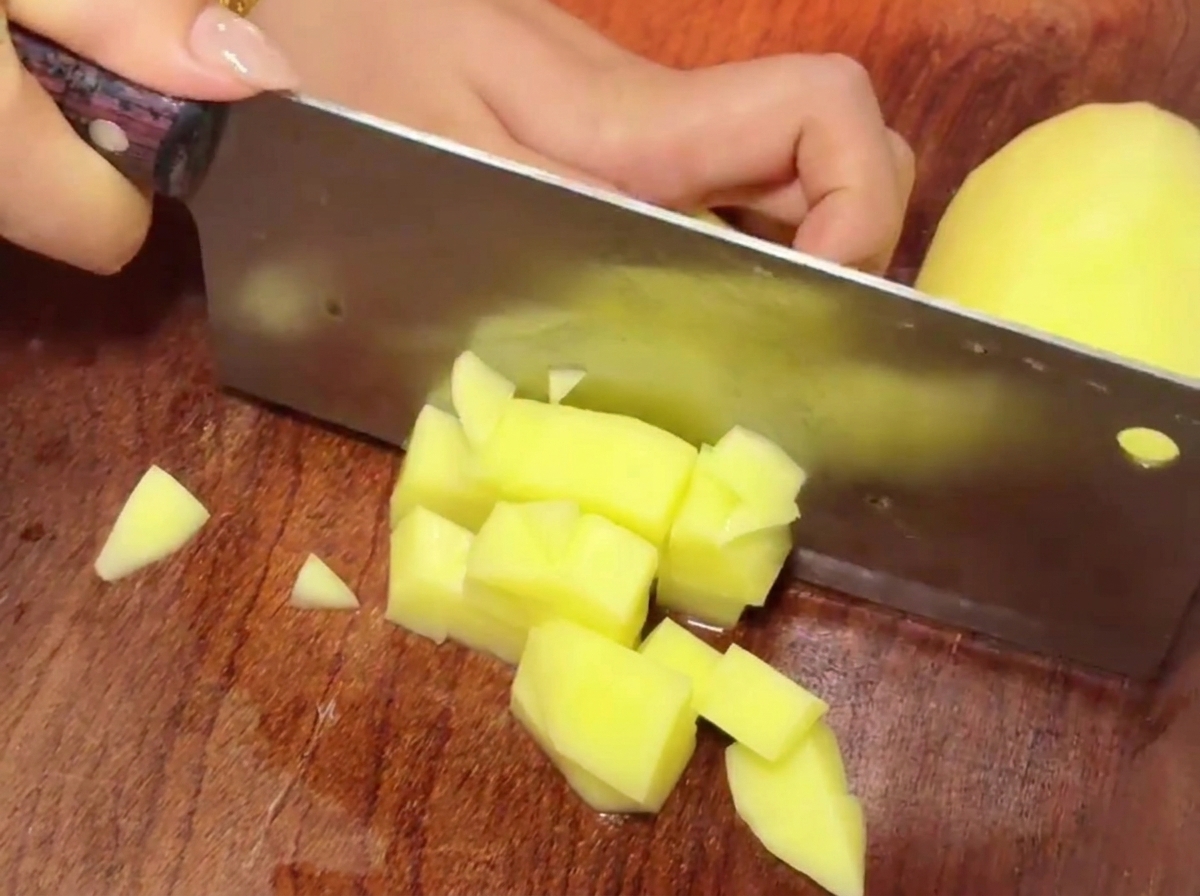 Un couperet coupant des morceaux de pommes de terre jaunes sur une planche à découper en bois pour former de petits cubes.