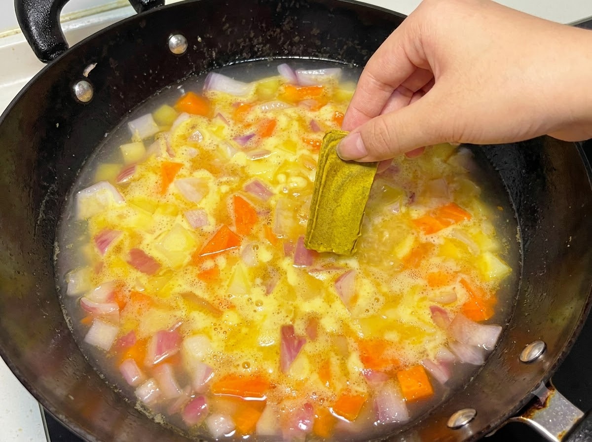Une main plaçant un bloc de curry solide dans une casserole deau et de légumes frémissante.