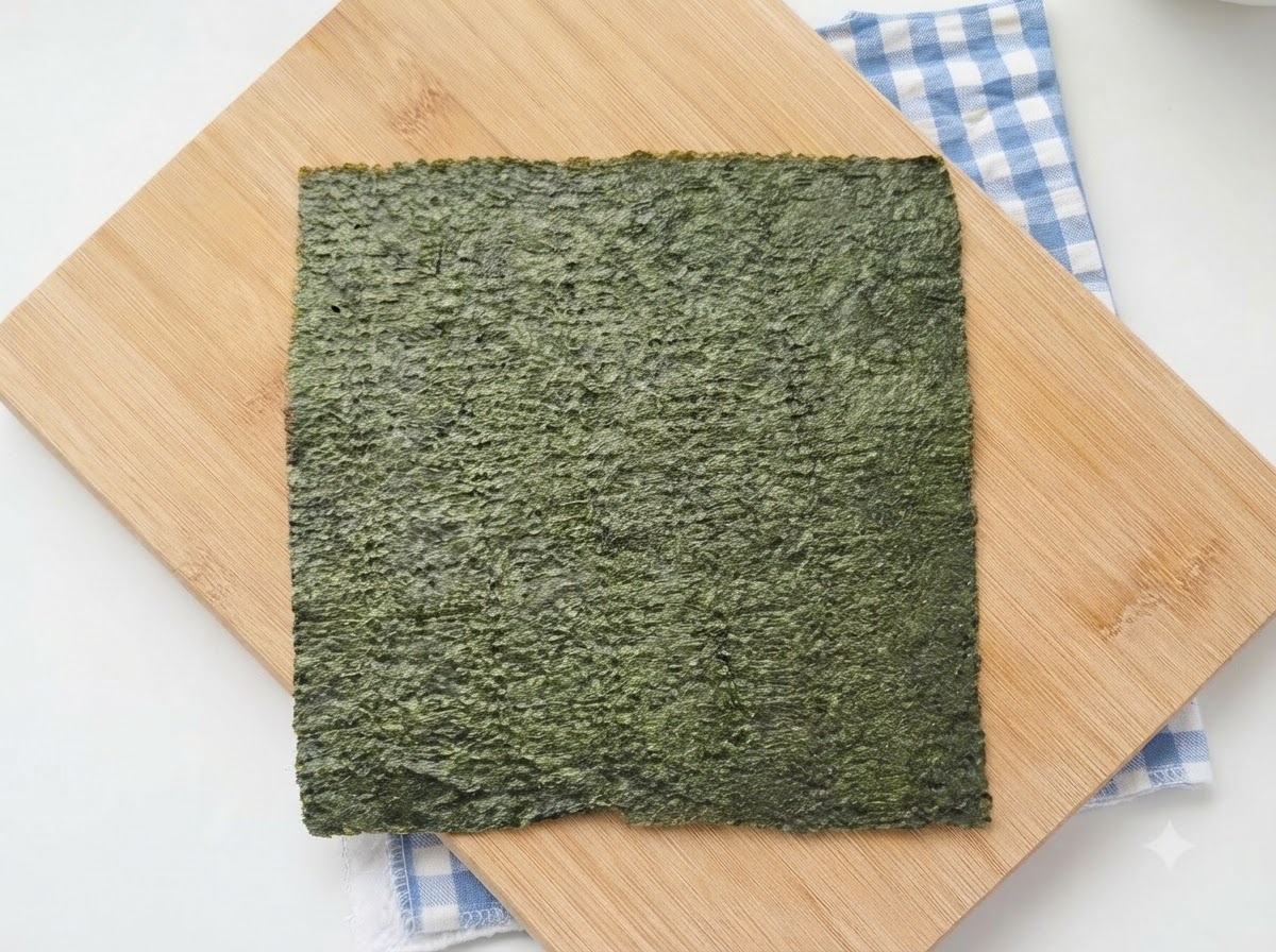 Une feuille entière de nori vert posée sur une planche à découper en bois avec un tissu à carreaux bleus en dessous.