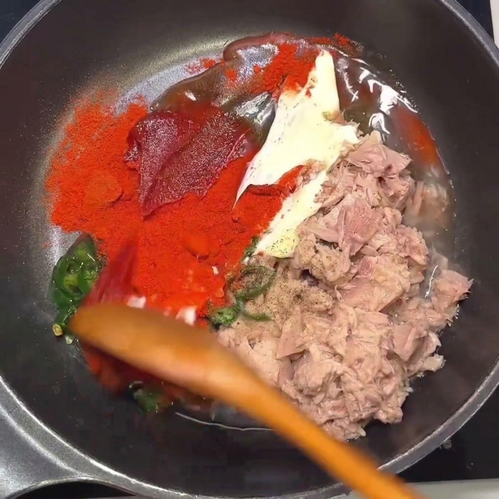 Une cuillère en bois mélangeant le thon, le piment et les assaisonnements dans une poêle.