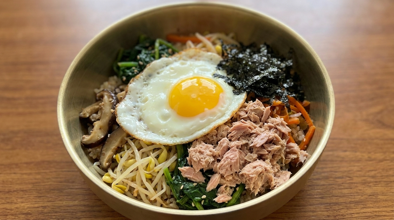 Un bol coloré de bibimbap au thon comprenant du riz assaisonné, des légumes frais, des champignons, des paillettes dalgues, du thon en boîte et un œuf au plat.