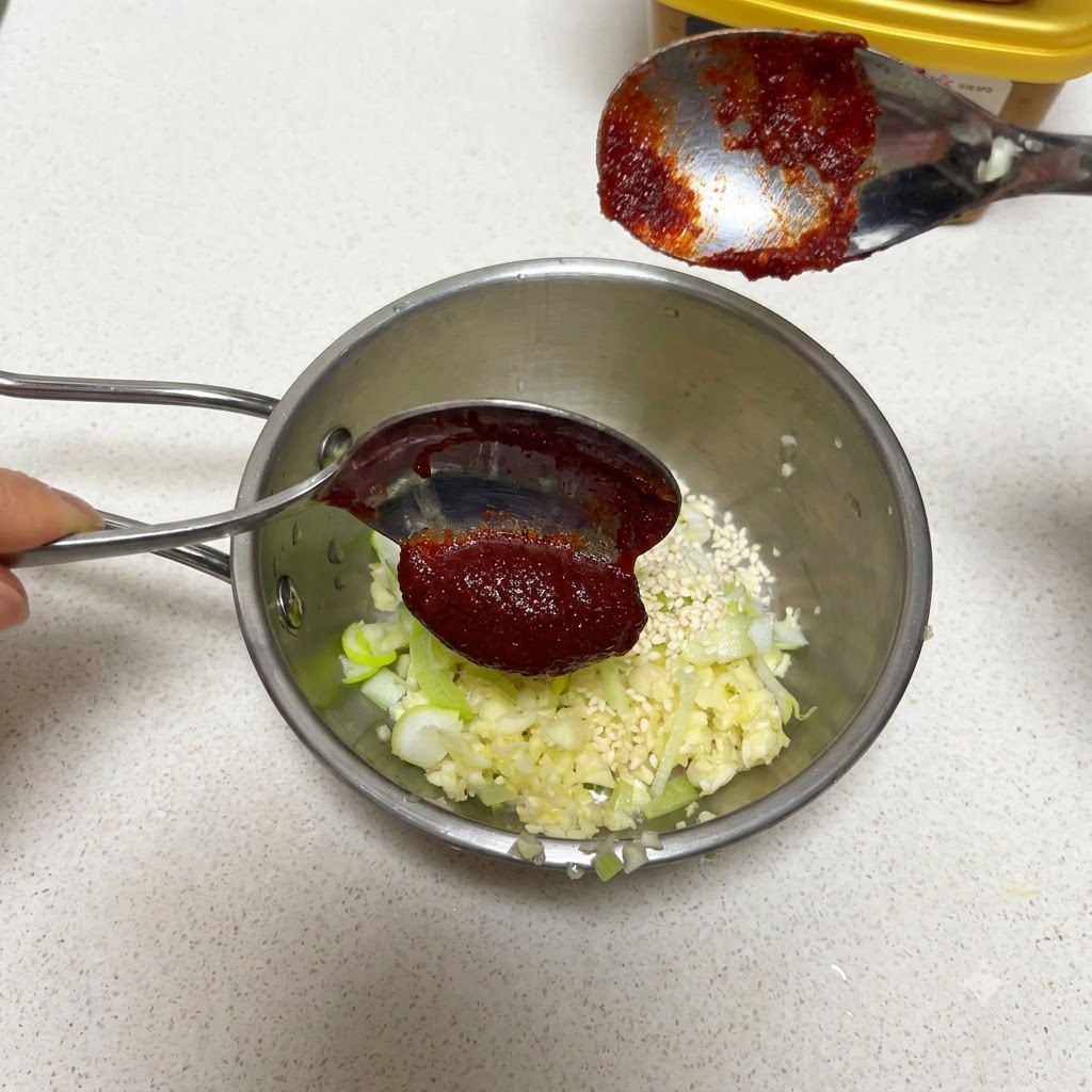 Une cuillère de pâte de piment coréenne tenue au-dessus dune poêle avec de lail et des graines de sésame.