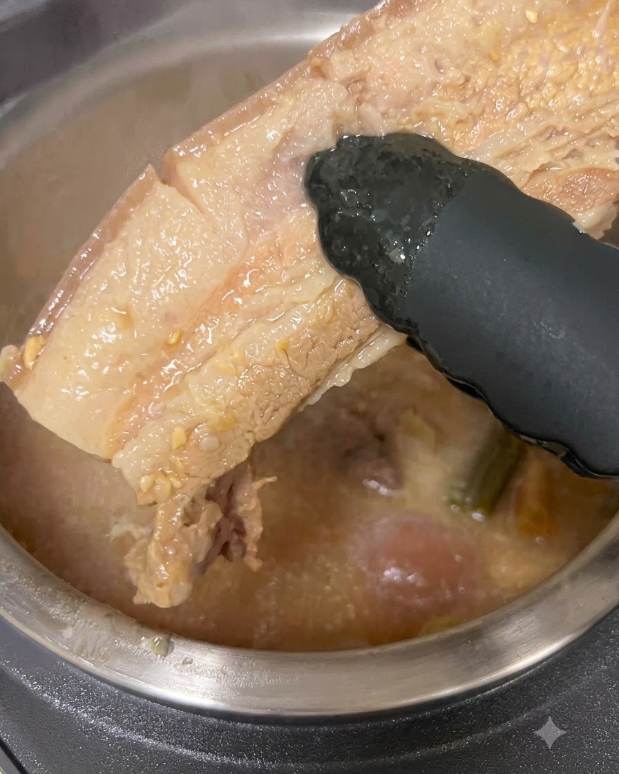 Un morceau épais de poitrine de porc cuite soulevé hors dun pot de bouillon avec des pinces noires.