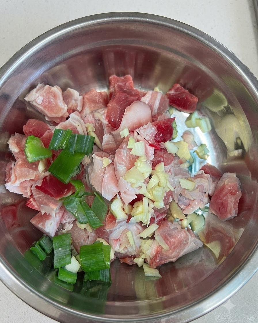 Morceaux de poitrine de porc crue dans un bol en métal, mélangés à de lail, des oignons verts et de la pâte de piment rouge.