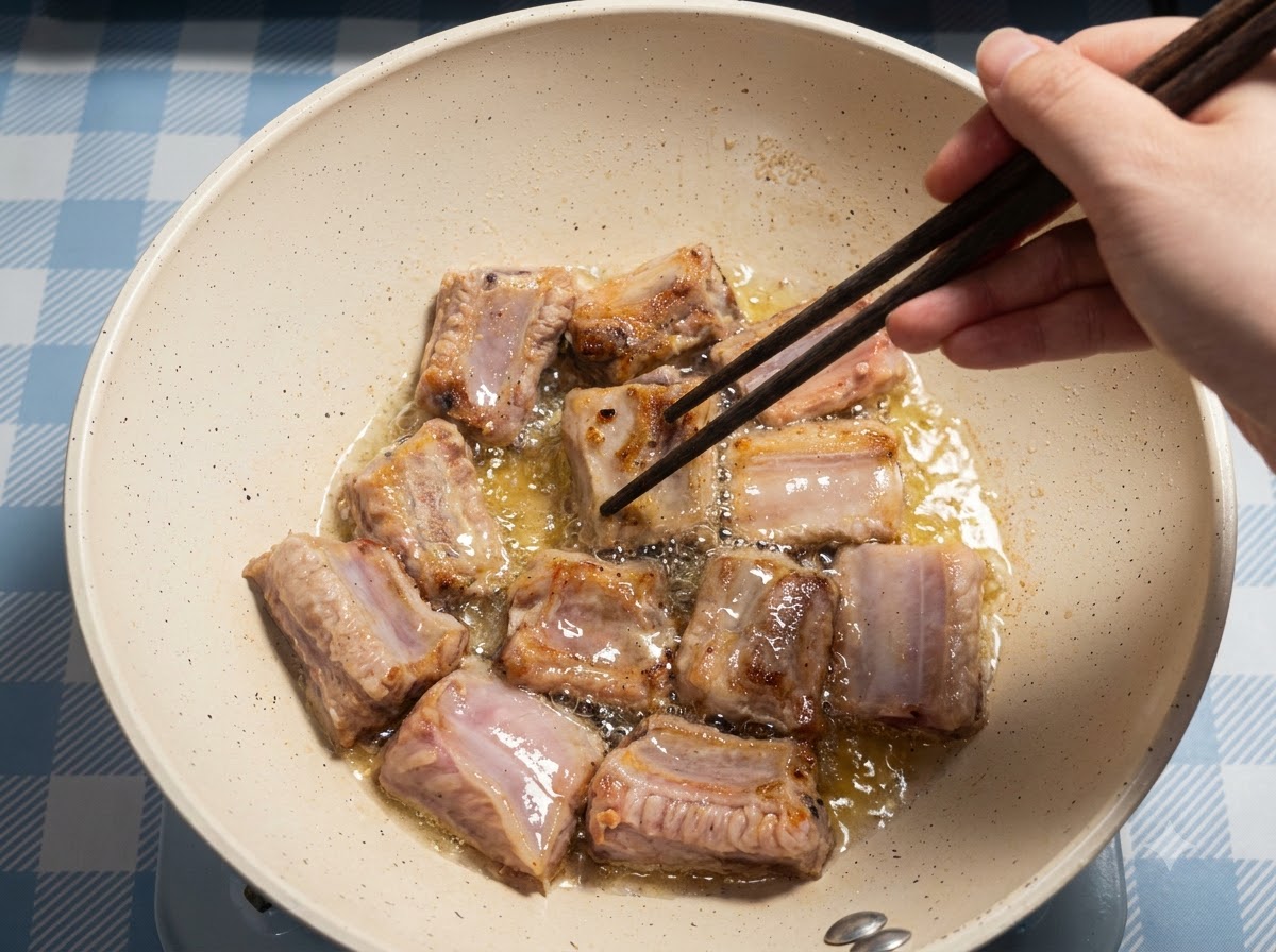 Plusieurs morceaux de travers de porc en train de frire dans une poêle, avec une main utilisant des baguettes pour les retourner.